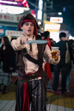 A person dressed as a pirate in a lively nighttime street setting, holding a plastic cup of beer and pointing a toy sword.