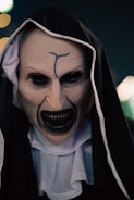 A person dressed in a dark, sinister costume resembling a nun with a menacing mask. The mask features exaggerated, eerie facial features with sharp teeth and a dark hood covering the head. The expression is haunting and unsettling.