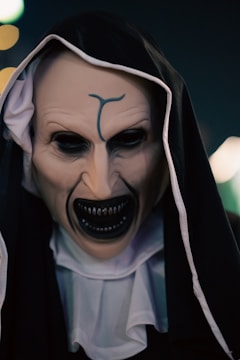 A person dressed in a dark, sinister costume resembling a nun with a menacing mask. The mask features exaggerated, eerie facial features with sharp teeth and a dark hood covering the head. The expression is haunting and unsettling.