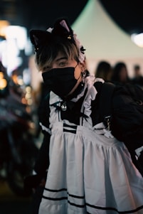 A person dressed in a black and white maid outfit, complete with a headband featuring cat ears. They are wearing a black face mask and standing in a dimly lit area. The background has blurred lights and silhouettes, indicating a night-time setting with possibly a street festival or fair atmosphere.