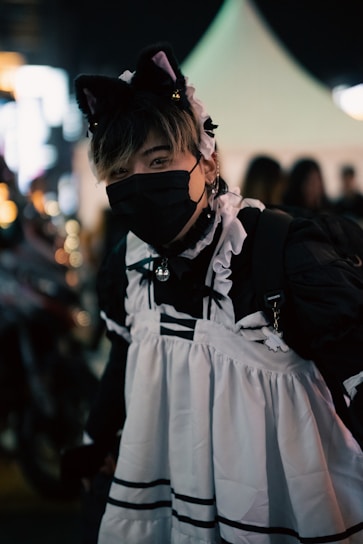 A person dressed in a black and white maid outfit, complete with a headband featuring cat ears. They are wearing a black face mask and standing in a dimly lit area. The background has blurred lights and silhouettes, indicating a night-time setting with possibly a street festival or fair atmosphere.