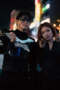 Two individuals dressed in dark, tactical costumes stand confidently in a nighttime urban setting. One holds a prominent weapon, while the other has short, red hair and holds a small object, possibly a weapon as well. A sense of determination is evident on their faces, complemented by dramatic lighting and blurred city lights in the background.