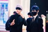 Two individuals are dressed in costumes, possibly cosplaying characters from an anime or comic. One person has silver hair and is wearing a black eyemask, along with a black outfit, while signaling with their hand. The other person has dark blue hair, a black face mask, and is dressed in a suit with a white shirt and tie. The background features several people and appears to be in an indoor, possibly public setting with bright lighting and blurred details.