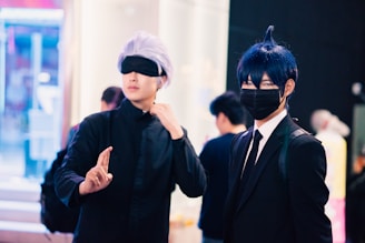Two individuals are dressed in costumes, possibly cosplaying characters from an anime or comic. One person has silver hair and is wearing a black eyemask, along with a black outfit, while signaling with their hand. The other person has dark blue hair, a black face mask, and is dressed in a suit with a white shirt and tie. The background features several people and appears to be in an indoor, possibly public setting with bright lighting and blurred details.