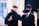 Two individuals are dressed in costumes, possibly cosplaying characters from an anime or comic. One person has silver hair and is wearing a black eyemask, along with a black outfit, while signaling with their hand. The other person has dark blue hair, a black face mask, and is dressed in a suit with a white shirt and tie. The background features several people and appears to be in an indoor, possibly public setting with bright lighting and blurred details.
