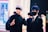 Two individuals are dressed in costumes, possibly cosplaying characters from an anime or comic. One person has silver hair and is wearing a black eyemask, along with a black outfit, while signaling with their hand. The other person has dark blue hair, a black face mask, and is dressed in a suit with a white shirt and tie. The background features several people and appears to be in an indoor, possibly public setting with bright lighting and blurred details.