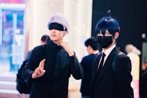 Two individuals are dressed in costumes, possibly cosplaying characters from an anime or comic. One person has silver hair and is wearing a black eyemask, along with a black outfit, while signaling with their hand. The other person has dark blue hair, a black face mask, and is dressed in a suit with a white shirt and tie. The background features several people and appears to be in an indoor, possibly public setting with bright lighting and blurred details.