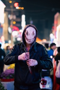 A person wearing a hooded jacket and a mask with a sinister smile holds a large knife with what appears to be blood on it. The background is an urban nightlife scene with colorful blurred lights and people.