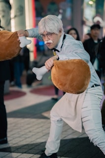A person dressed in a costume consisting of white hair, glasses, and a mustache, holding large plush drumsticks. The setting seems to be urban, possibly during a public event or cosplay gathering. The atmosphere is lively with onlookers in the background.