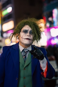 A person is dressed in a costume resembling a popular comic book character with green hair and white face paint, featuring dark eye makeup and a red smile painted onto the face. They wear a blue suit jacket, green vest, and a tie with a patterned design. The background is a blurred cityscape with colorful lights visible.