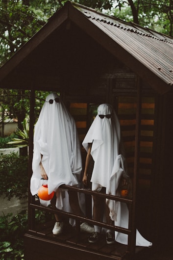 Two figures dressed in ghost costumes made from white sheets stand side by side on a wooden porch. Both figures are holding orange pumpkin-shaped containers, typically used for collecting treats during Halloween. The porch is surrounded by greenery, and there is a sense of a dim, overcast day.
