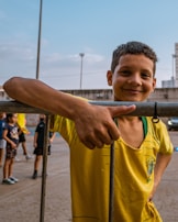 A smiling boy wearing a yellow sports jersey leans casually against a metal fence. In the background, other children are gathered on what appears to be a sports court. The sky is clear and blue, with some buildings and a light pole visible.