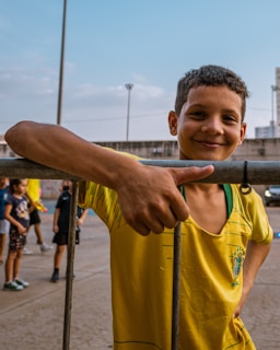 A smiling boy wearing a yellow sports jersey leans casually against a metal fence. In the background, other children are gathered on what appears to be a sports court. The sky is clear and blue, with some buildings and a light pole visible.