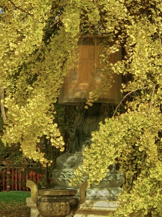 A serene statue of a figure is partially obscured by vibrant golden foliage, creating an enchanting scene. An ornate, round vessel with intricate designs is placed in front of the statue. The background features dense greenery and a glimpse of a wooden structure.