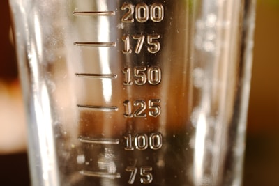A close-up view of a transparent measuring container with measurement markings ranging from 75 to 200 milliliters. The surface of the container appears reflective with some light and shadow play.