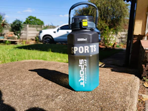 A large 2000ml sports water bottle with a gradient design from black on top to blue at the bottom, placed on a concrete surface in an outdoor setting. The bottle has a handle and a yellow latch. In the background, there is a white vehicle, a grassy lawn, and some garden plants.