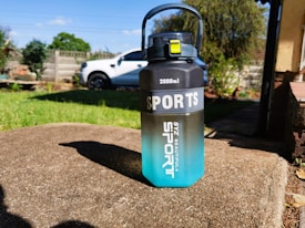 A large 2000ml sports water bottle with a gradient design from black on top to blue at the bottom, placed on a concrete surface in an outdoor setting. The bottle has a handle and a yellow latch. In the background, there is a white vehicle, a grassy lawn, and some garden plants.