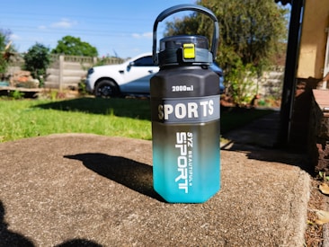 A large 2000ml sports water bottle with a gradient design from black on top to blue at the bottom, placed on a concrete surface in an outdoor setting. The bottle has a handle and a yellow latch. In the background, there is a white vehicle, a grassy lawn, and some garden plants.