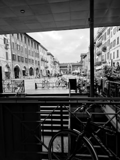 A monochrome photo captures a city street scene with old-style buildings featuring arched doorways and shuttered windows. The street is lined with bicycles, with people seen walking in the background. The foreground includes a covered area with railings, leading up to a bike resting against the structure. The sky is partially cloudy.