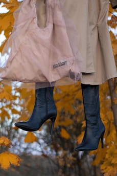 A pink bag with a sheer overlay and the word 'IGANNAIT' printed on it hangs alongside a pair of dark high-heeled boots. Both items are suspended against a backdrop of vibrant orange autumn leaves.