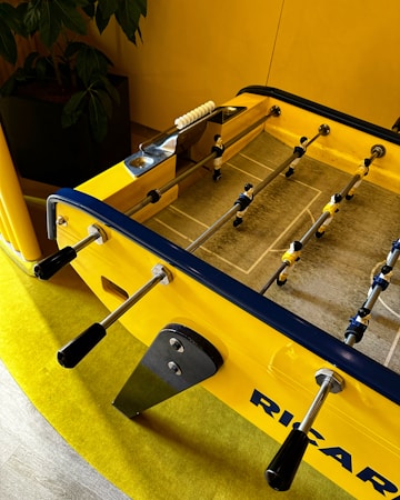 A brightly colored foosball table with yellow and blue accents is positioned on a yellow carpet. Metal rods with black handles run across the table, with small player figures attached. A leafy green plant is in the background next to a yellow wall.