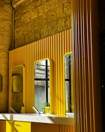 A modern bathroom with bright yellow paneling and mirrors on a textured stone wall. Below the mirrors, there is a white countertop with two sleek faucets and a transparent soap dispenser. A partially visible window allows light to cast shadows across the surfaces.