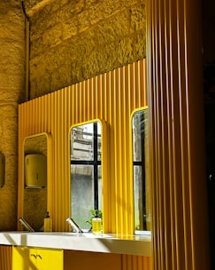 A modern bathroom with bright yellow paneling and mirrors on a textured stone wall. Below the mirrors, there is a white countertop with two sleek faucets and a transparent soap dispenser. A partially visible window allows light to cast shadows across the surfaces.