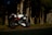A sleek, modern motorcycle is parked on the roadside near a dense forest. The bike is primarily white with red accents and blue-tinted wheels. Tall trees with textured bark are visible in the background, casting shadows over the road.