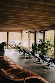 A peaceful indoor seating area with wooden lounge chairs and metal-framed chairs. Large glass windows surround the space, providing a view of lush greenery outside. The ceiling features exposed wooden beams, adding to the natural ambiance. Several potted plants are arranged throughout the area, enhancing the tranquil and cozy atmosphere.