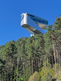 A modern architectural structure with a futuristic design is elevated on a pedestal above a dense, green forest. The building has a sleek, angular form and large tinted windows, set against a clear, blue sky.
