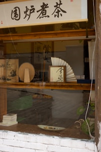 A shop window display features various traditional objects, including Asian calligraphy scrolls, a decorative fan, and some books. The setting has a rustic appearance with wooden frames and a white brick wall. A green plant adds to the natural ambiance of the scene.