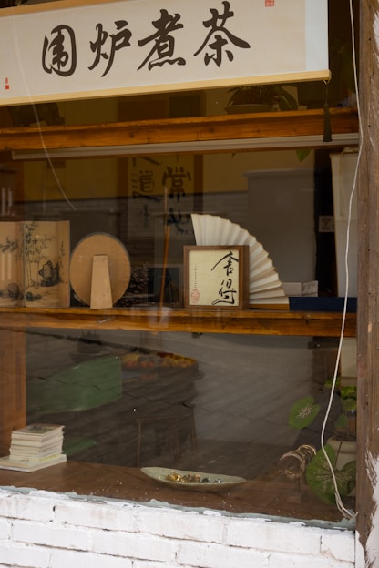 A shop window display features various traditional objects, including Asian calligraphy scrolls, a decorative fan, and some books. The setting has a rustic appearance with wooden frames and a white brick wall. A green plant adds to the natural ambiance of the scene.