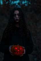 A person with long, curly hair, dressed in dark clothing, holds a carved pumpkin with a glowing red inside. The background is dim and forested, creating a mysterious and eerie atmosphere.