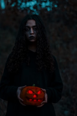 A person with long, curly hair, dressed in dark clothing, holds a carved pumpkin with a glowing red inside. The background is dim and forested, creating a mysterious and eerie atmosphere.