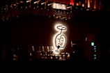 A dimly lit bar area is highlighted by a glowing, stylized wall art depicting a wine glass with a face. Racks of hanging upside-down glasses are visible above, and a counter is layered with various glassware. Soft, warm lighting creates a cozy, intimate atmosphere.