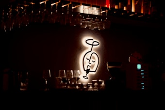 A dimly lit bar area is highlighted by a glowing, stylized wall art depicting a wine glass with a face. Racks of hanging upside-down glasses are visible above, and a counter is layered with various glassware. Soft, warm lighting creates a cozy, intimate atmosphere.