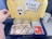 An airline meal tray with various food items including two plastic containers with food, a bread roll, a foil-wrapped main dish, a pack of wet wipes, and a sachet with meal information. The background shows a colorful packaging lid featuring a cartoon panda chef and airplane graphics.