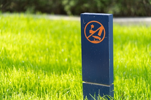 A blue post with an orange sign is placed on a vibrant green lawn. The sign features a crossed-out dog symbol, indicating a restriction on dog walking.