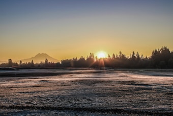 The image depicts a serene landscape at sunrise with the sun rising just above the tree line, casting rays across a frosty or dewy field. A distant mountain is visible on the horizon, adding depth to the background.