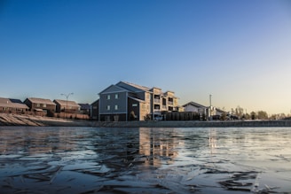 A residential area with houses and an apartment building situated by a frozen body of water. The ice on the water has a textured surface. The sky is clear and displays a gradient from blue to light yellow, indicating either sunrise or sunset.