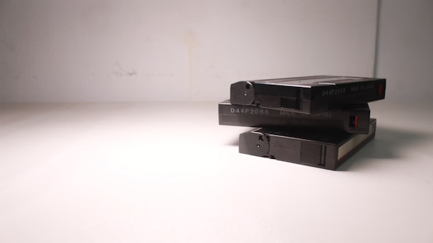 A stack of three VHS tapes placed on a plain, light-colored surface. The tapes are marked with a code and 'Made in Japan' text. The composition is simple, with minimalistic lighting casting soft shadows.
