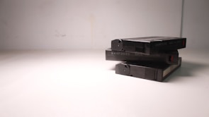 A stack of three VHS tapes placed on a plain, light-colored surface. The tapes are marked with a code and 'Made in Japan' text. The composition is simple, with minimalistic lighting casting soft shadows.