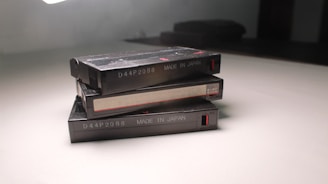 Three vintage cassette tapes are stacked on a white surface. Each cassette has labeling that reads 'D44P2088 MADE IN JAPAN,' and there are red accents near the edges. The lighting casts a shadow on the smooth surface.