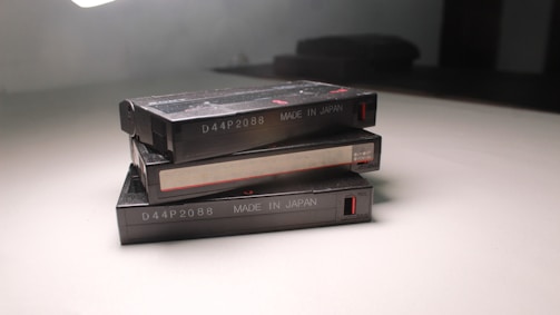 Three vintage cassette tapes are stacked on a white surface. Each cassette has labeling that reads 'D44P2088 MADE IN JAPAN,' and there are red accents near the edges. The lighting casts a shadow on the smooth surface.