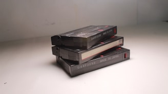 Three compact cassette tapes are stacked on top of each other against a plain neutral background. Each cassette is black with a red label and white printed text indicating 'MADE IN JAPAN' and a code number. The surface of the tapes reflects a bit of light, suggesting a slightly glossy finish.
