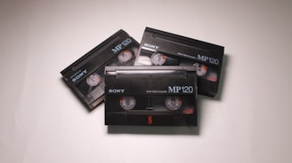 Three black Sony MP120 video cassettes are arranged on a light surface. Each cassette has visible reels and orange details.