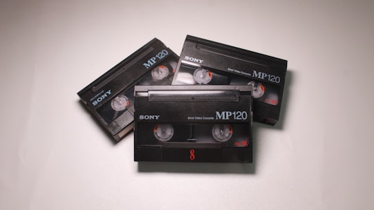 Three black Sony MP120 video cassettes are arranged on a light surface. Each cassette has visible reels and orange details.
