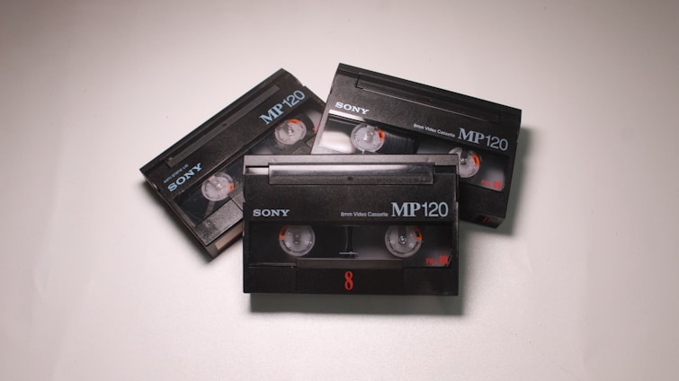 Three black Sony MP120 video cassettes are arranged on a light surface. Each cassette has visible reels and orange details.