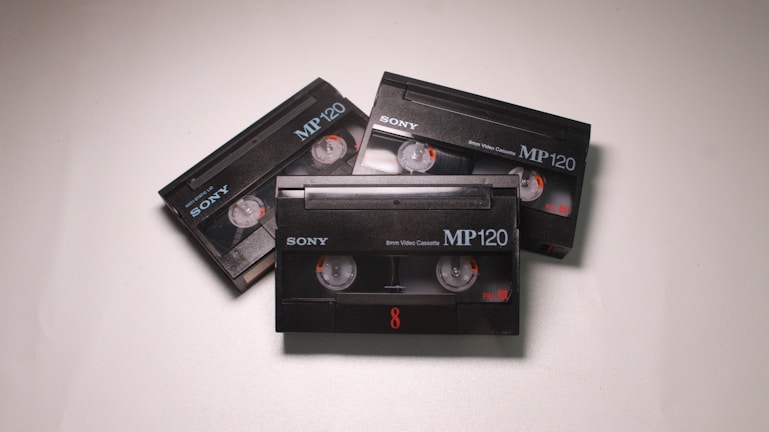 Three black Sony MP120 video cassettes are arranged on a light surface. Each cassette has visible reels and orange details.