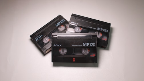 Three black Sony MP120 video cassettes are arranged on a light surface. Each cassette has visible reels and orange details.